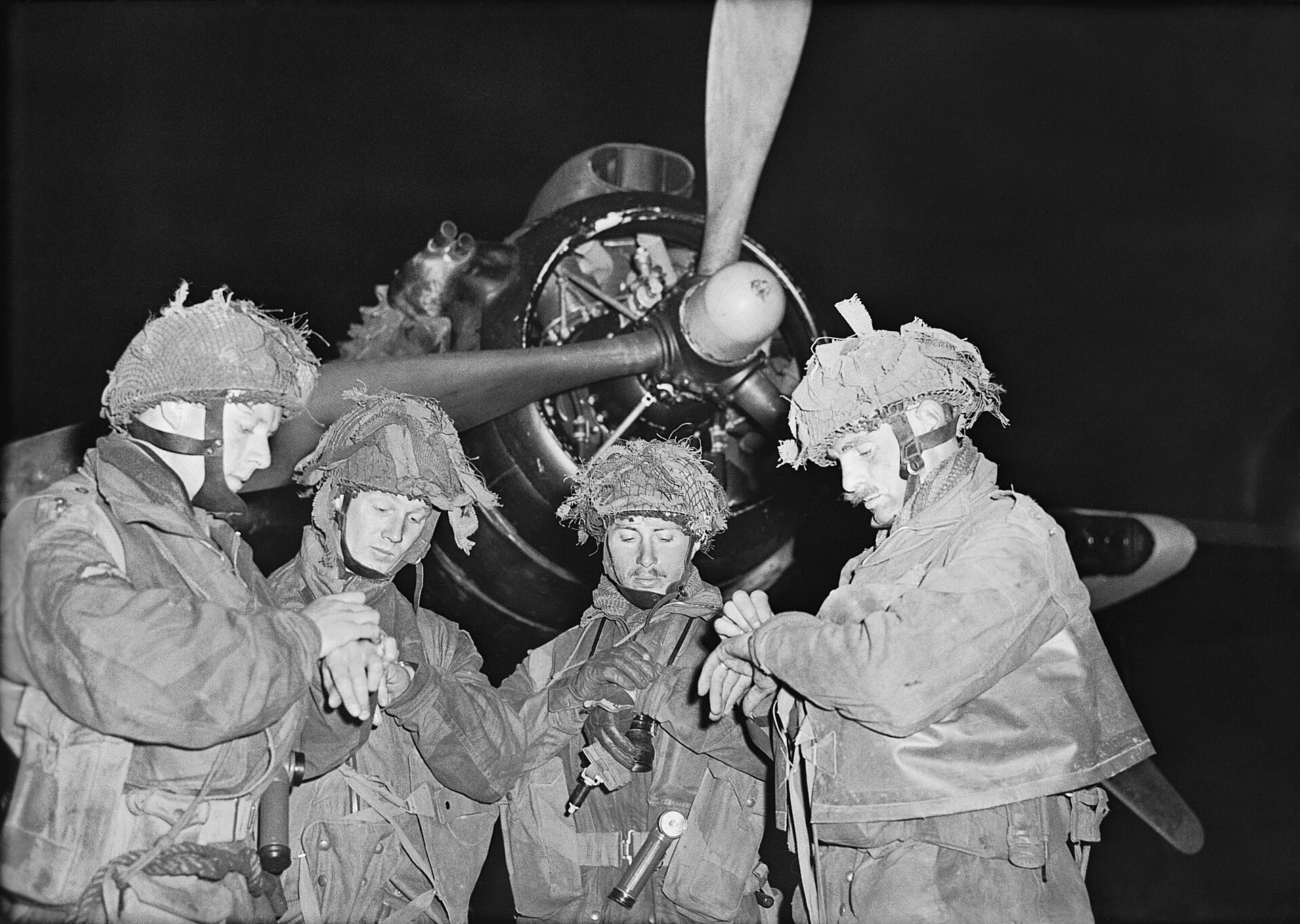 Four British paratroopers in helmets checking their wristwatches in front of an aircraft propeller at night, 5 June 1944