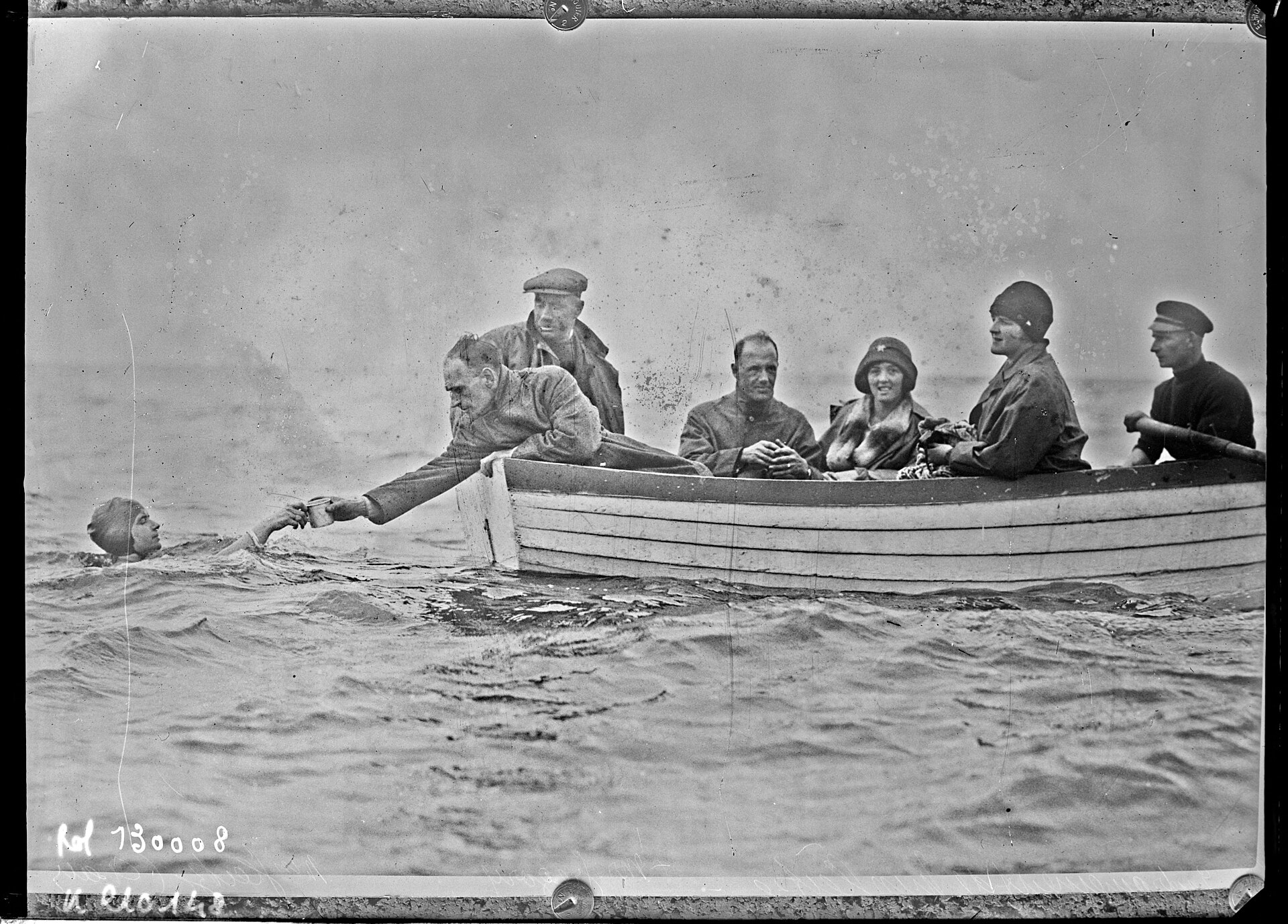 A 1920s press photograph of Mercedes Gleitze swimming beside a rowing boat while a man leans over to hand her a cup